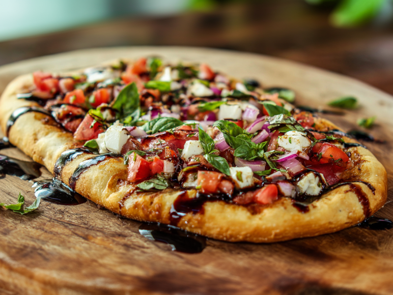 Eine Bruschetta-Pizza auf einem Holzuntersetzer. Sie ist mit etwas Balsamico-Creme beträufelt. Im Hintergrund erkennt man leicht angeschnittene Kräuter.