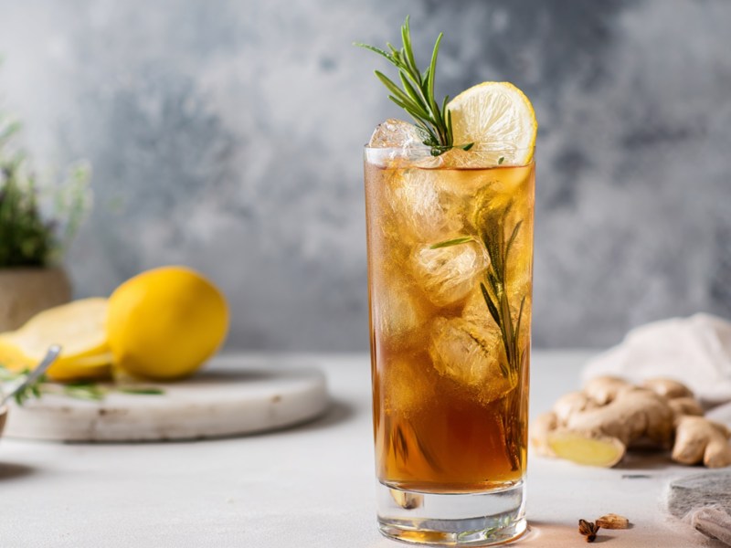 Ein Glas Earl Grey Ginger Spritz, der mit einer Scheibe Zitrone und einem Zweig Rosmarin garniert ist. Im Hintergrund kann man Zitronen und Ingwerwurzeln, genauso wie einen Topf mit frischem Rosmarin erkennen.