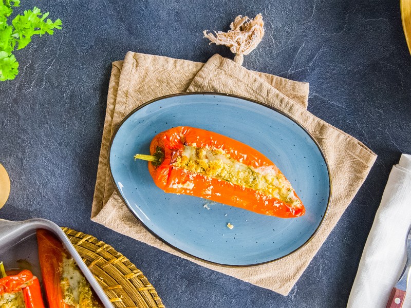 Ein blauer Teller mit einer gefüllten Paprika mit Feta und Kräutern. Ringsum kann man eine Stoffserviette mit Besteckt, frische Kräuter, Paprika und einen Salzstreuer erkennen.