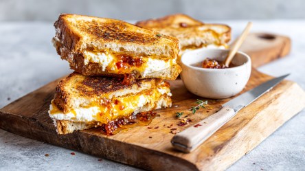 Ein aufgeschnittenes Grilled Cheese Sandwich mit Ziegenkäse, Honig und Chili, das auf einem Holzbrett auf einer Arbeitsplatte liegt. Auf dem Brett steht zudem eine kleine Schale mit Chili-Marmelade. Auch ein Messer liegt dabei.