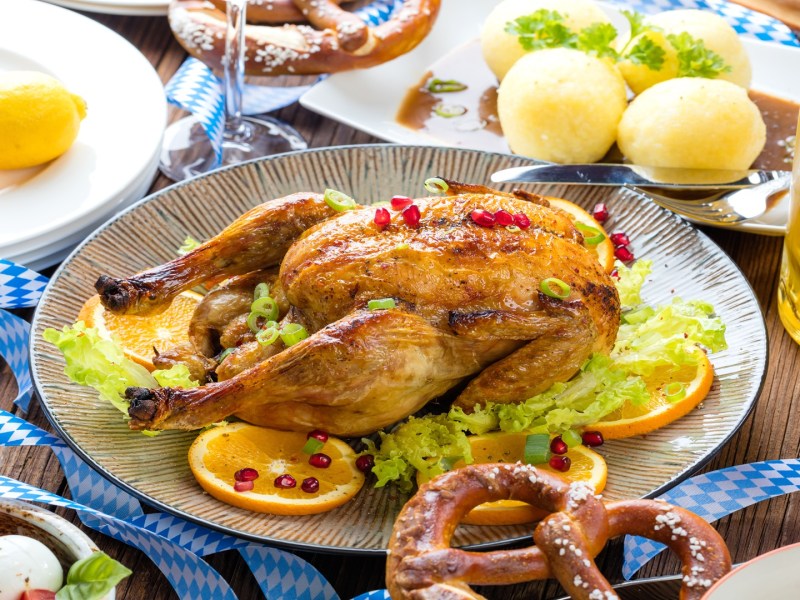 Ein Wiesnhendl auf einem Teller. Es ist umgeben von Orangenscheiben, Granatapfelkernen, Bier, Knödeln und Brezeln.