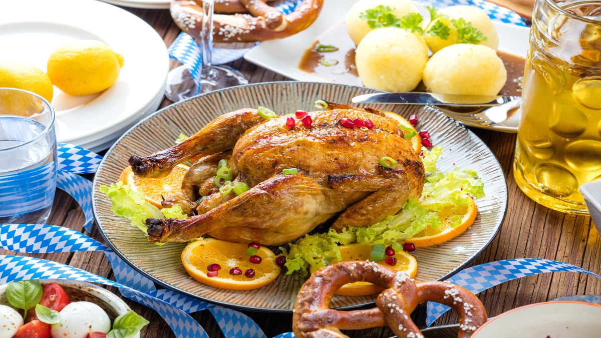 Ein Wiesnhendl auf einem Teller. Es ist umgeben von Orangenscheiben, Granatapfelkernen, Bier, Knödeln und Brezeln.