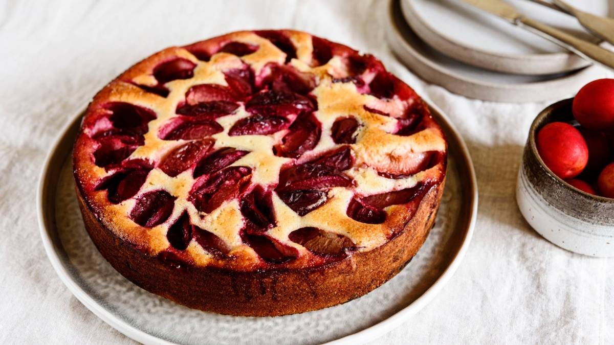 Ein ganzer Käsekuchen mit Pflaumen, der auf einem großen Teller platziert ist. Rechts daneben steht eine Schale mit Pflaumen. Im Hintergrund kann man Teller und Besteck erkennen.