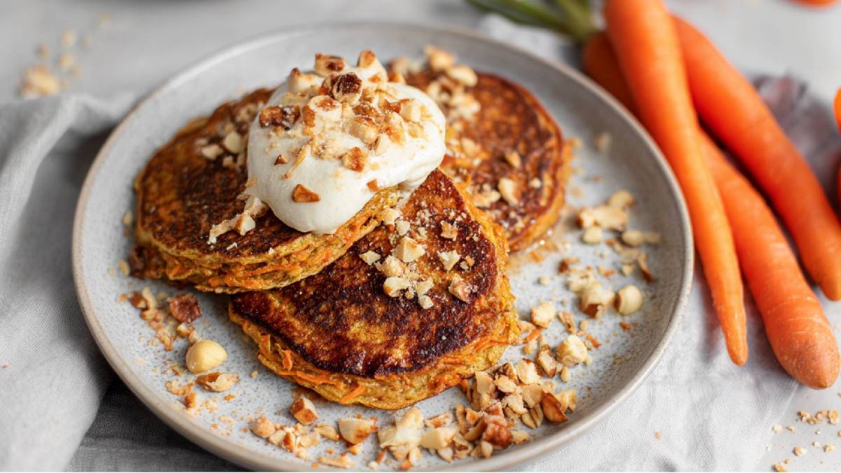 Drei Karotten-Haferflocken Pancakes auf einem Teller mit Joghurtklecks und Haselnüssen bestreut, frische Karotten daneben