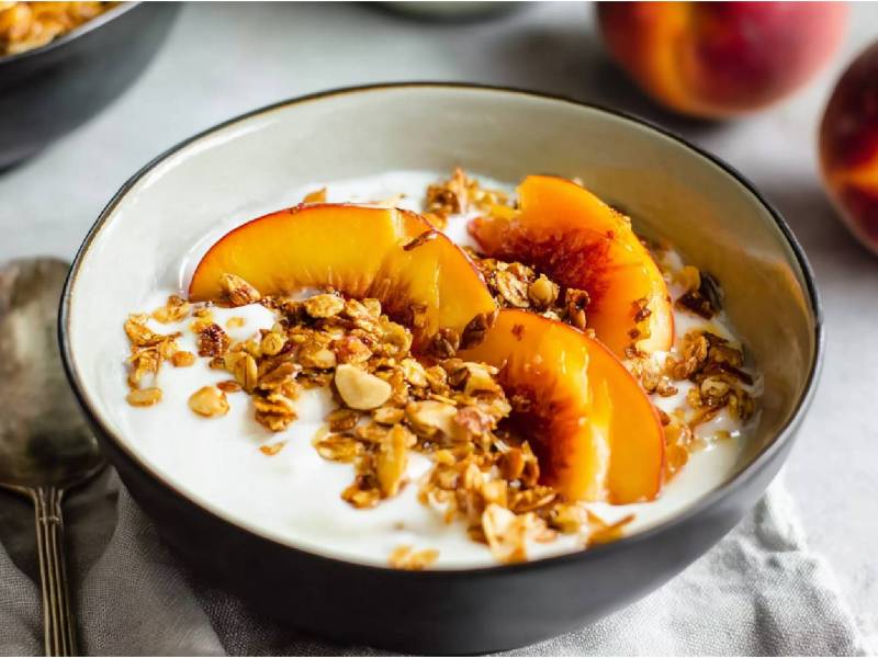 Kefir-Joghurt-Bowl mit gerösteten Pfirsichen auf einem Tisch, Pfirsiche im Hintergrund