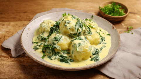 Mehre Knödel mir Käse-Spinat-Soße auf einem grauen Teller. Im Hintergrund ist noch ein Schälchen mit frischen Kräutern.