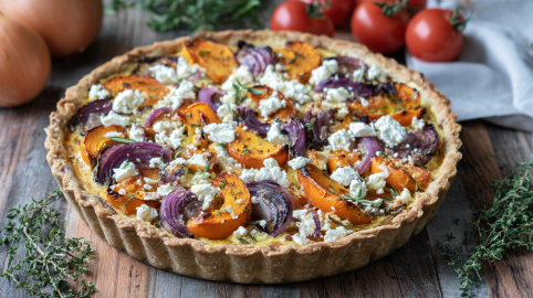 Eine Kürbis-Tomaten-Quiche steht auf einem Holztisch. Im Hintergrund links liegen zwei große Zwiebeln, rechts daneben ein paar Kräuter sowie Tomtaten.