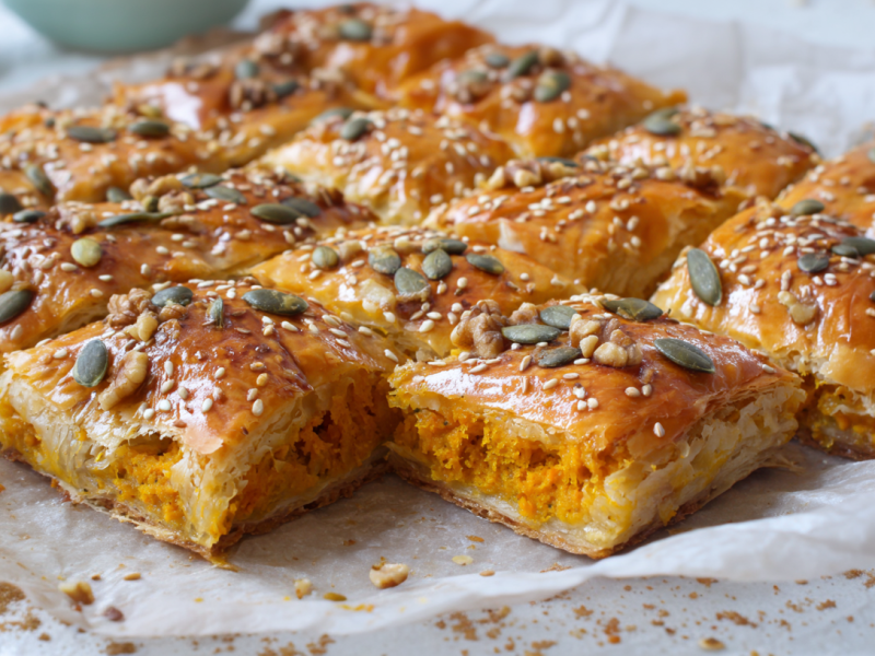 Ein, in Stücke geschnittener Kürbiskuchen vom Blech. Er ist mit gehackten Walnusskernen, Kürbiskernen und Sesam bestreut. Im Hintergrund sind zwei kleine Schalen zu sehen.