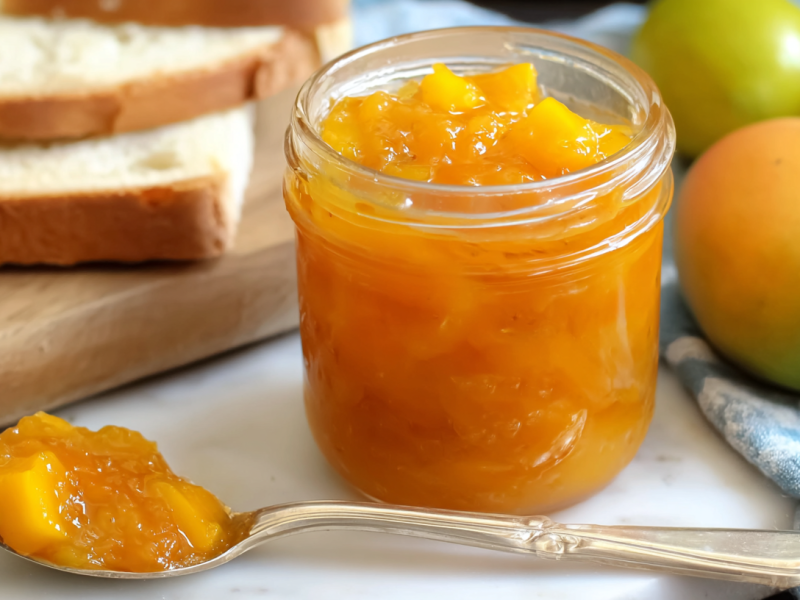 Ein Glas Mango-Limetten-Marmelade mit einem Löffel mit der Marmelade daneben.