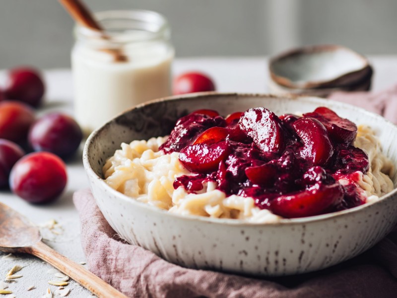 Ein Teller voll Milchnudeln mit Pflaumenkompott. Im Hintergrund steht ein Glas Milch und es liegen einige frische Pflaumen herum. Neben dem Teller liegt ein Holzlöffel.