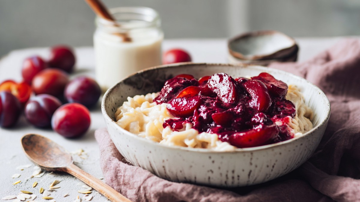 Ein Teller voll Milchnudeln mit Pflaumenkompott. Im Hintergrund steht ein Glas Milch und es liegen einige frische Pflaumen herum. Neben dem Teller liegt ein Holzlöffel.