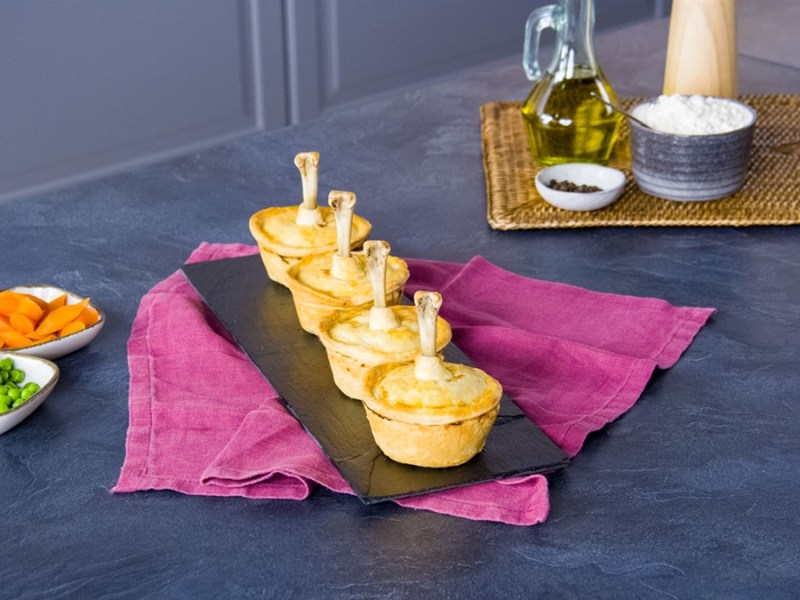 Vier Mini-Hähnchen-Pies sind auf einem Schieferbrett angerichtet. Daneben liegen noch Schalen mit Möhren und Erbsen. Im Hintergrund steht eine Flasche Öl sowie Pfeffer, Mehl und eine Pfeffermühle.