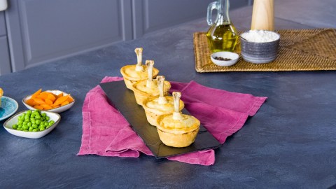 Vier Mini-Hähnchen-Pies sind auf einem Schieferbrett angerichtet. Daneben liegen noch Schalen mit Möhren und Erbsen. Im Hintergrund steht eine Flasche Öl sowie Pfeffer, Mehl und eine Pfeffermühle.