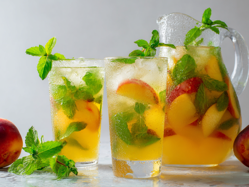 Zwei Gläser mit Nektarinen-Minz-Limonade. Daneben steht eine Glaskaraffe ebenfalls mit der Limo. Links und rechts im Bild liegen zwei Nektarinen. Vor den Gläsern liegt etwas Minze.