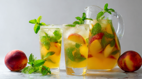 Zwei Gläser mit Nektarinen-Minz-Limonade. Daneben steht eine Glaskaraffe ebenfalls mit der Limo. Links und rechts im Bild liegen zwei Nektarinen. Vor den Gläsern liegt etwas Minze.