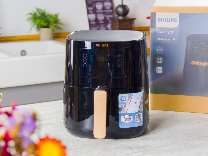 Ein Philips Airfryer 500 aus der Connected Series, der auf einer hellen Arbeitsplatte in einer Küche steht. Hinter dem Airfryer ist der Karton des Produkts platziert. Im Vordergrund kann man verschwommen einige bunte Blumen erkennen. Ansonsten kann man etwas vom Rest der Küche sehen.