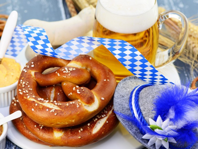 Brez'n, Obazda und weitere Oktoberfest-Klassiker liegen neben einer Maß Bier und einem typisch bayerischen Hut.