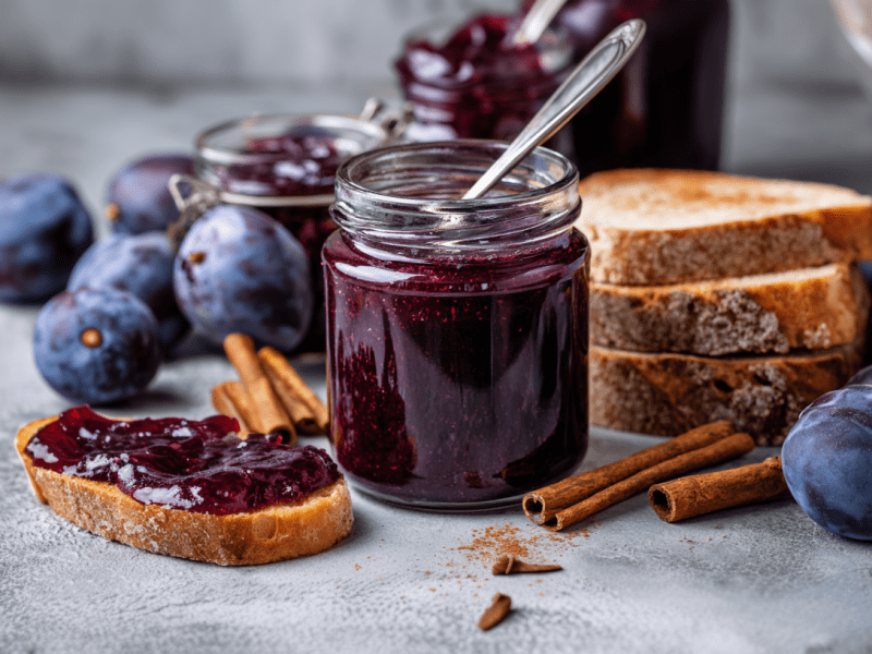 Einige Gläser mit Pflaumenmarmelade auf einem grauen Untergrund. Vor dem vordesten liegt ein Brot mit der Marmelade darauf. Drumherum sind einige Pflaumen sowie Zimtstangen verteilt.