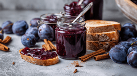 Einige Gläser mit Pflaumenmarmelade auf einem grauen Untergrund. Vor dem vordesten liegt ein Brot mit der Marmelade darauf. Drumherum sind einige Pflaumen sowie Zimtstangen verteilt.