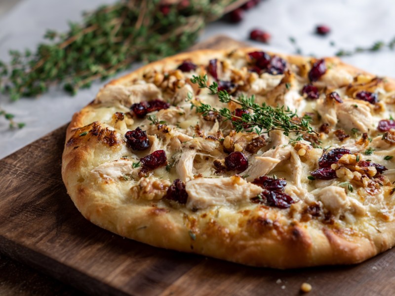 Ein Pizza mit Hähnchen, Brie und Cranberrys, die mit ein bisschen frischem Thymian garniert ist. Die Pizza lieg auf einem großen dunklen Holzbrett. Im Hintergrund liegen weitere Zweige Thymian und ein paar frische Cranberrys.