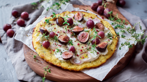Eine Polenta-Pizza mit Feigen und Trauben auf einem Holzuntersetzer. Die Pizza ist mit frischen Kräutern garniert. Im Hintergrund sieht man noch weitere Trauben liegen.