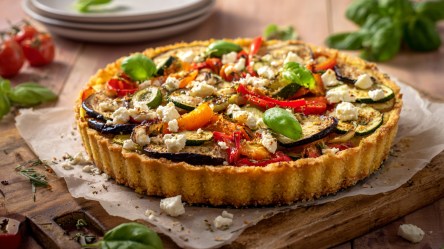 Eine Polenta-Tarte mit Gemüse auf einem Holzbrett mit Backpapier. Im Hintergrund kann man einen Stapel mit Tellern. Außerdem liegen einige frische Zutaten herum.