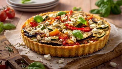 Eine Polenta-Tarte mit Gemüse auf einem Holzbrett mit Backpapier. Im Hintergrund kann man einen Stapel mit Tellern. Außerdem liegen einige frische Zutaten herum.