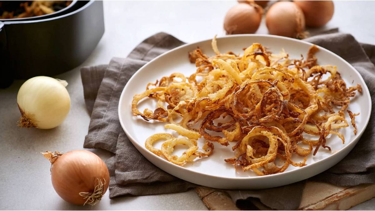 Schale mit Röstzwiebeln aus dem Airfryer, frische Zwiebeln und Airfryer im Hintergrund