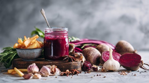Ein Glas mit selbst gemachtem Rote-Bete-Ketchup, das auf einem Holzbrett steht. Daneben steht eine kleine Schale mit Pommes. Ringsum liegen Knoblauch, frische Rote Bete sowie Gewürze herum.