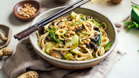 Eine Shiitake-Nudelpfanne, angerichtet in einer Schüssel. Darauf liegen zwei Essstäbchen. Die Schüssel steht auf einem Tuch. Drumherum liegen einige Erdnüsse verteilt.