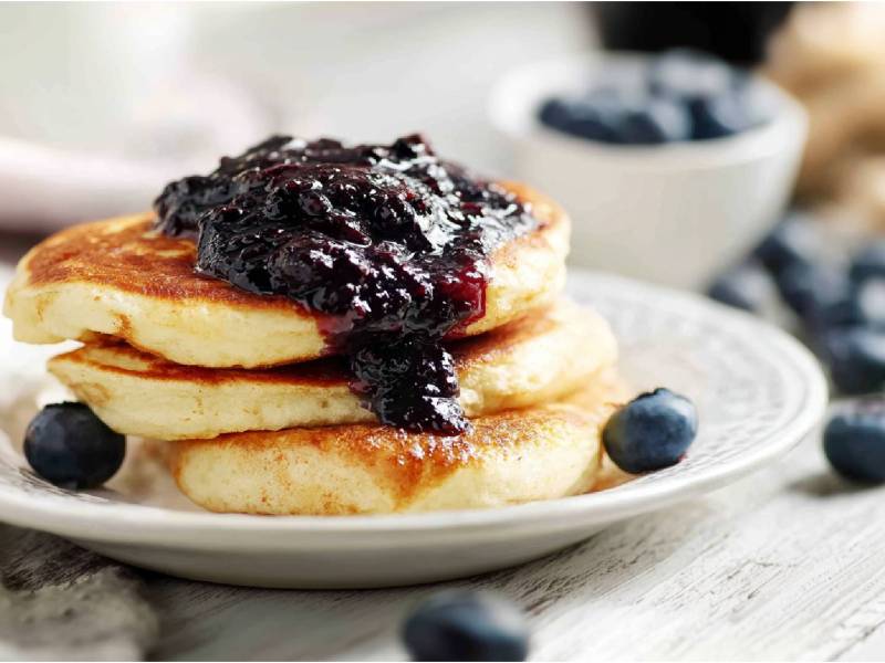 Teller mit Skyr-Pancakes mit Blåbärssylt und Blaubeeren in einer Schale dahinter