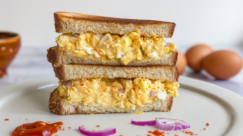 Ein Spiegeleiersalat-Sandwich, dessen beiden Hälften aufeinander gestapelt auf einem Teller liegen. Auf dem Teller ist ein Klecks Sriracha-Soße und liegen ein paar Zwiebeln. Im Hintergrund kann man Eier sehen.