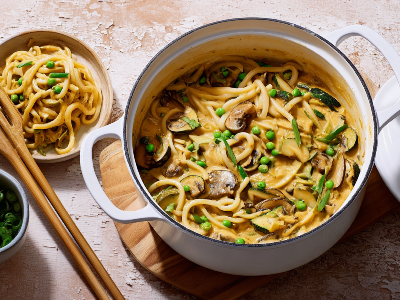 Ein Topf mit Udon-Nudeln-One-Pot ist zu sehen. Daneben steht eine kleine Schüssel mit dem Gericht und ein Schälchen mit Frühlingszwiebeln.