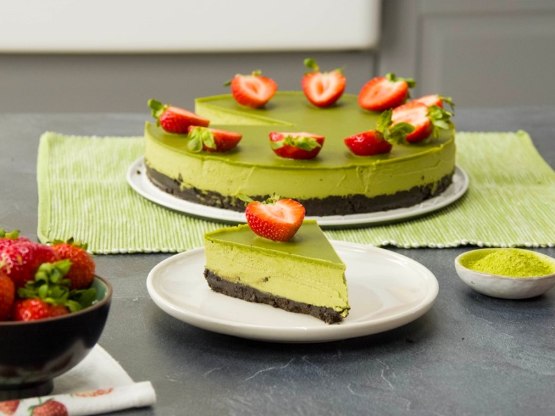 Ein Stück vegane Matcha-Torte auf einem Teller. Im Hintergrund steht die gesamte, mit Erdbeeren dekorierte Torte. Rechts neben dem kleinen Teller ist ein Schälchen Matcha-Pulver.