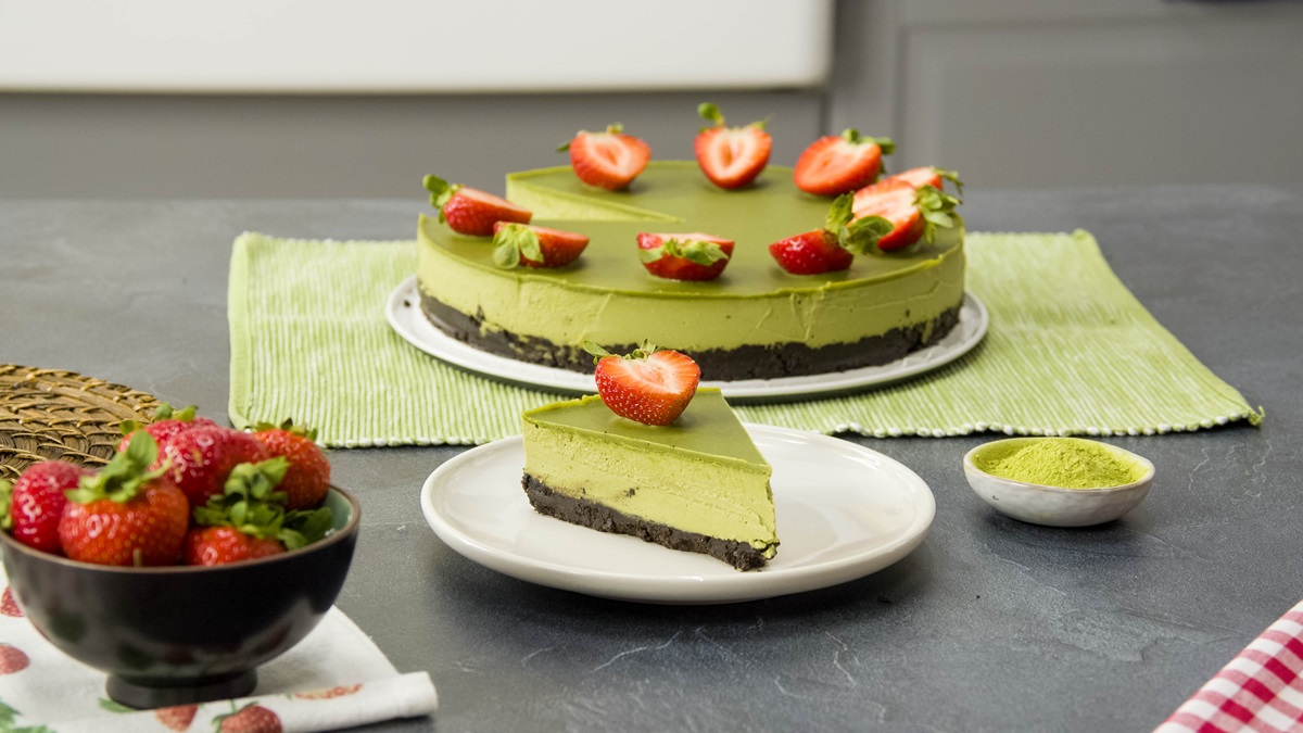 Ein Stück vegane Matcha-Torte auf einem Teller. Im Hintergrund steht die gesamte, mit Erdbeeren dekorierte Torte. Rechts neben dem kleinen Teller ist ein Schälchen Matcha-Pulver.
