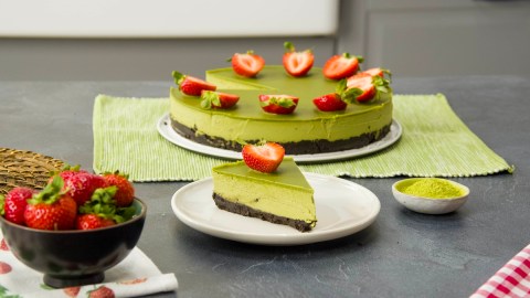 Ein Stück vegane Matcha-Torte auf einem Teller. Im Hintergrund steht die gesamte, mit Erdbeeren dekorierte Torte. Rechts neben dem kleinen Teller ist ein Schälchen Matcha-Pulver.