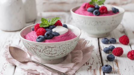 Zwei Schüsseln mit Vispipuuro auf einem weißen Holztisch. Neben den Schüsseln liegen frische Beeren. Vor der vorderen Schüssel liegt ein Löffel.