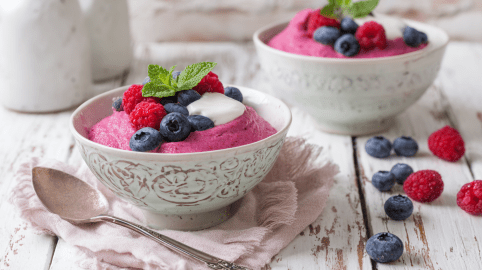 Zwei Schüsseln mit Vispipuuro auf einem weißen Holztisch. Neben den Schüsseln liegen frische Beeren. Vor der vorderen Schüssel liegt ein Löffel.