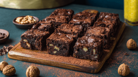 Ein Brett mit zwölf Walnuss-Pflaumen-Brownies. Sie sind mit Kakaopulver bestäubt. Um das Brett herum liegen einige Walnüsse verteilt.
