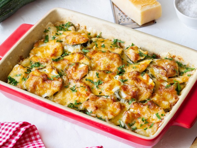 Zucchini-Auflauf in einer roten Auflaufform, garniert mit Schnittlauch. Daneben liegt ein Stück Parmesan.
