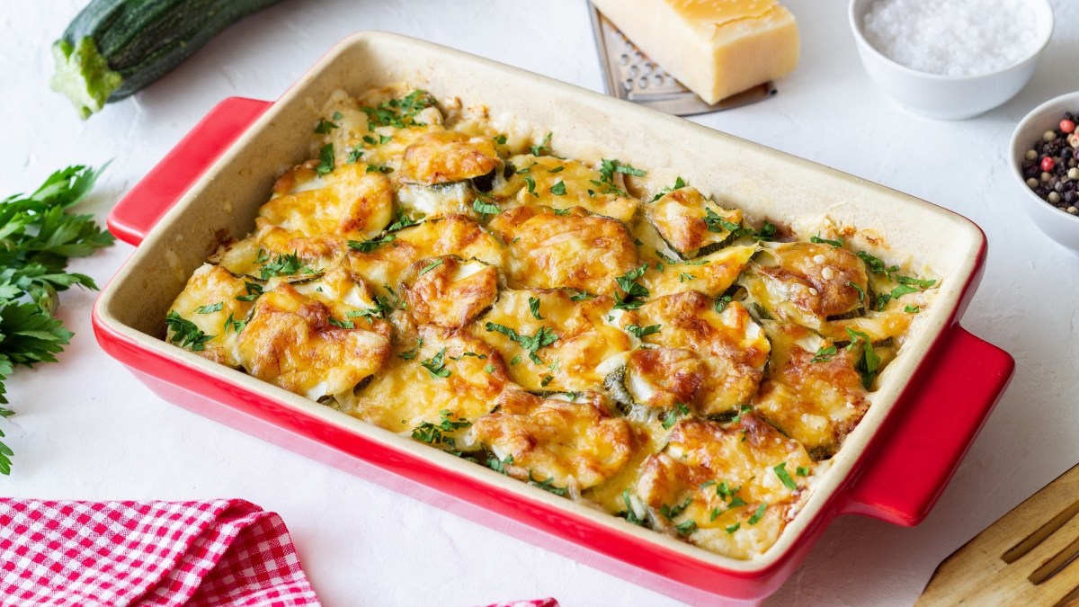 Zucchini-Auflauf in einer roten Auflaufform, garniert mit Schnittlauch. Daneben liegt ein Stück Parmesan.
