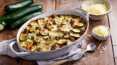 Zucchini-Hackfleisch-Gratin in einer Auflaufform, daneben liegen Zucchini und etwas Käse.