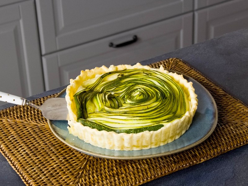 Eine Zucchini-Ricotta-Tarte auf einem grauen Teller auf einer Küchenmatte. Die Tarte ist angeschnitten und ein Kuchenheber liegt bereit, sie zu servieren.