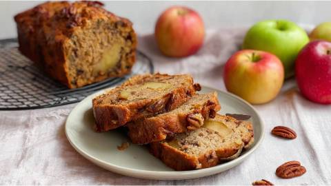 Drei Scheiben Apfel-Pekannuss-Brot auf einem Teller, angeschnittenes Brot und Äpfel im Hintergrund