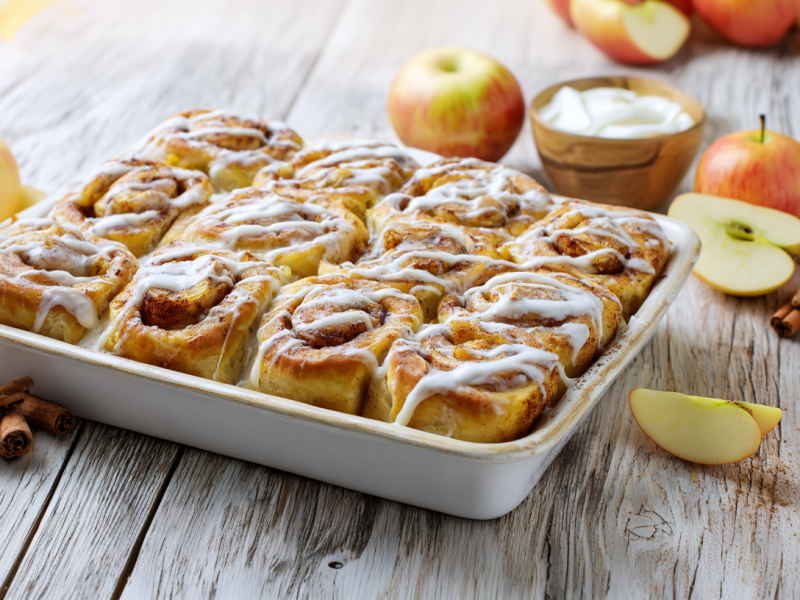 Ein Backblech voller Apfel-Puddingschnecken. Sie sind mit einem cremigen Frosting bestrichen. Um die Form herum liegen Äpfel und Zimtstangen verteilt.