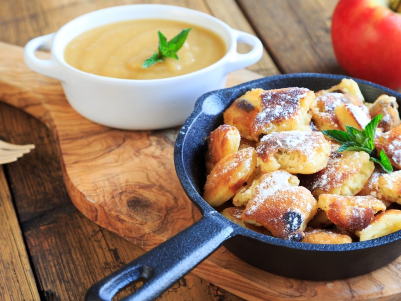 Apfelchmarrn in einer kleinen Pfanne. Daneben steht eine Schale mit Apfelmus und es liegt ein roter Apfel.