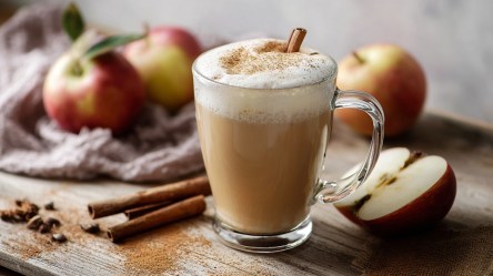 Ein Apple Cider Chai Latte, der auf einem Tisch steht. Daneben sind Zimtstangen, ein aufgeschnittener Apfel und ganz Äpfel zu sehen.