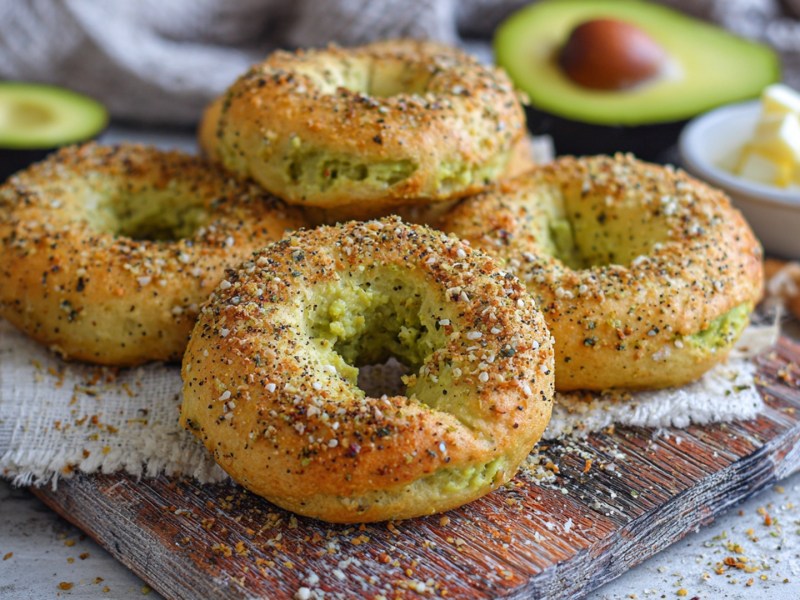 Einige Avocado-Bagel, die auf einem Holzbrett platziert sind. Ringsum liegen Avocadohälften sowie eine Schale mit Butterwürfeln ist zu sehen.