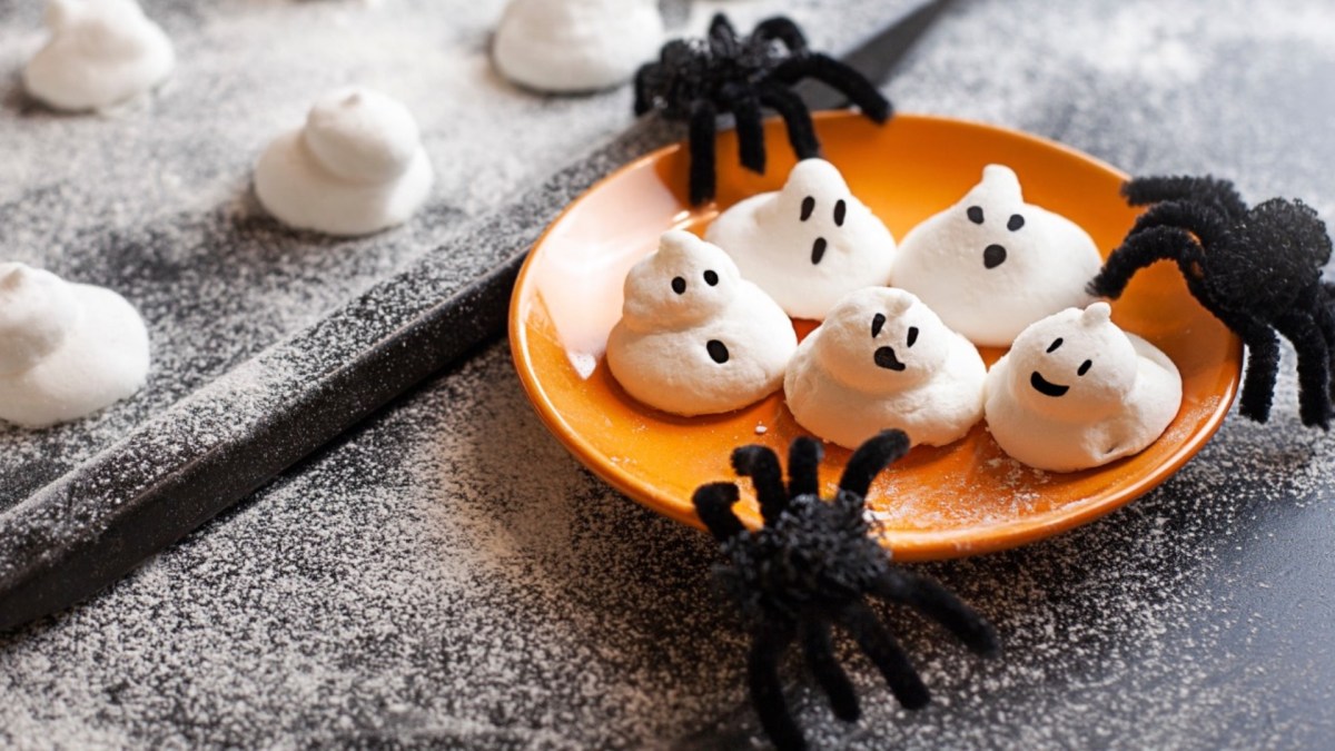 Fünf süße Baiser-Geister auf einem orangen Teller. Spinnen als Halloween-Deko liegen daneben.