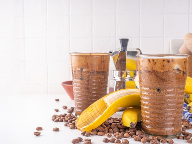 Zwei Gläser mit Bananen-Kaffee-Smoothie. Eine Banane und Kaffeebohnen liegen als Dekoration außen herum.